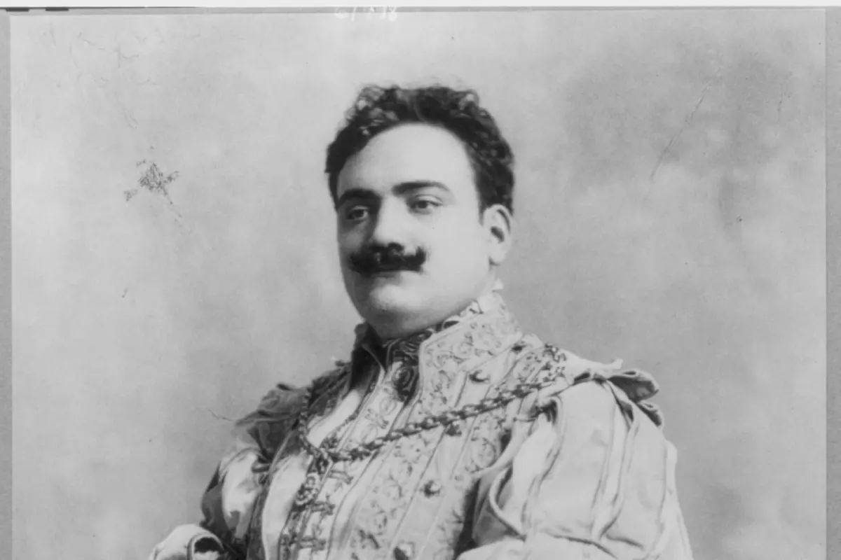 A black and white studio portrait of the tenor Enrico Caruso in a velvet and brocade stage costume, with a moustache and a plumed hat in his hand.