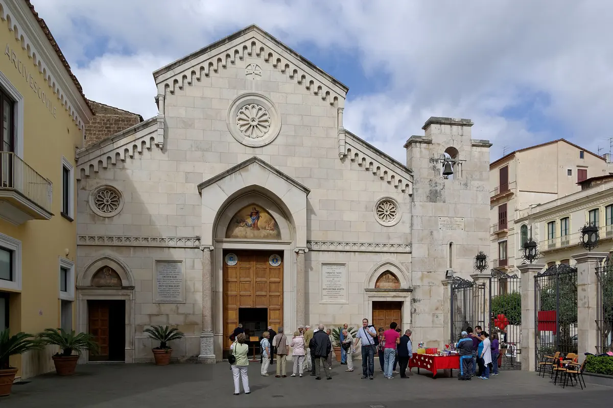 The pale stone front of the cathedral of Sorrento with a round window and a pointed doorway, with visitors gathered in the square in front.