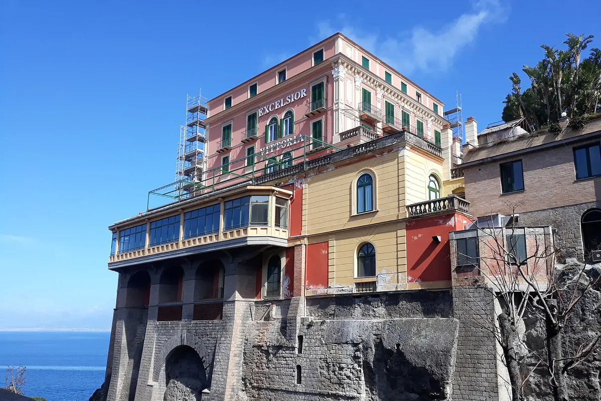 A pink and ochre hotel with green shutters and a glazed balcony standing on a high stone cliff above the sea in Sorrento.