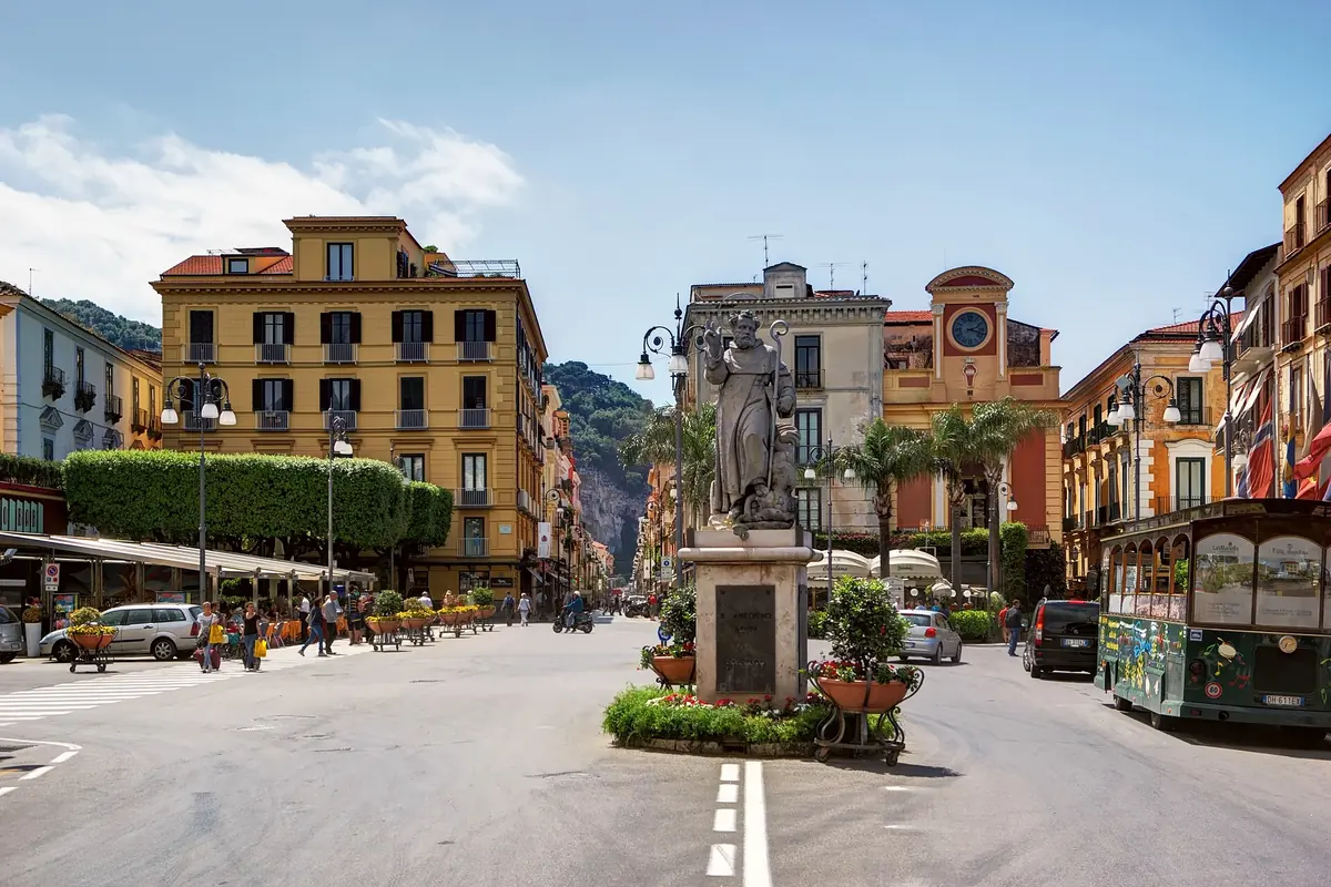 A wide square in Sorrento with ochre and terracotta buildings, a statue on a plinth in the middle and palm trees.